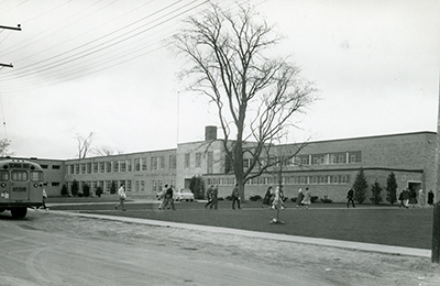 Old black and white picture of Dr. G.W. Williams school, with kids walking towards the school and an old school bus on the left