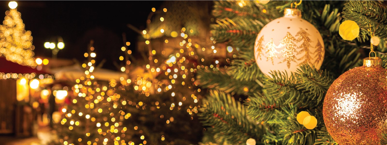 Closeup of Christmas ornaments on a tree and Christmas lights and another tree in the background