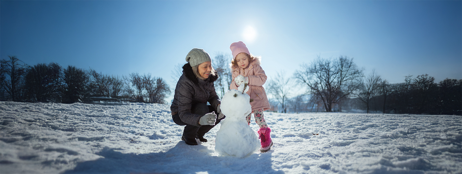 A woman and child building a snowman in the middle of a large snow field, with trees in the background