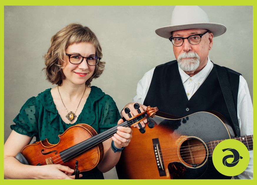 A woman holding a fiddle and a man holding a guitar looking directly at the camera