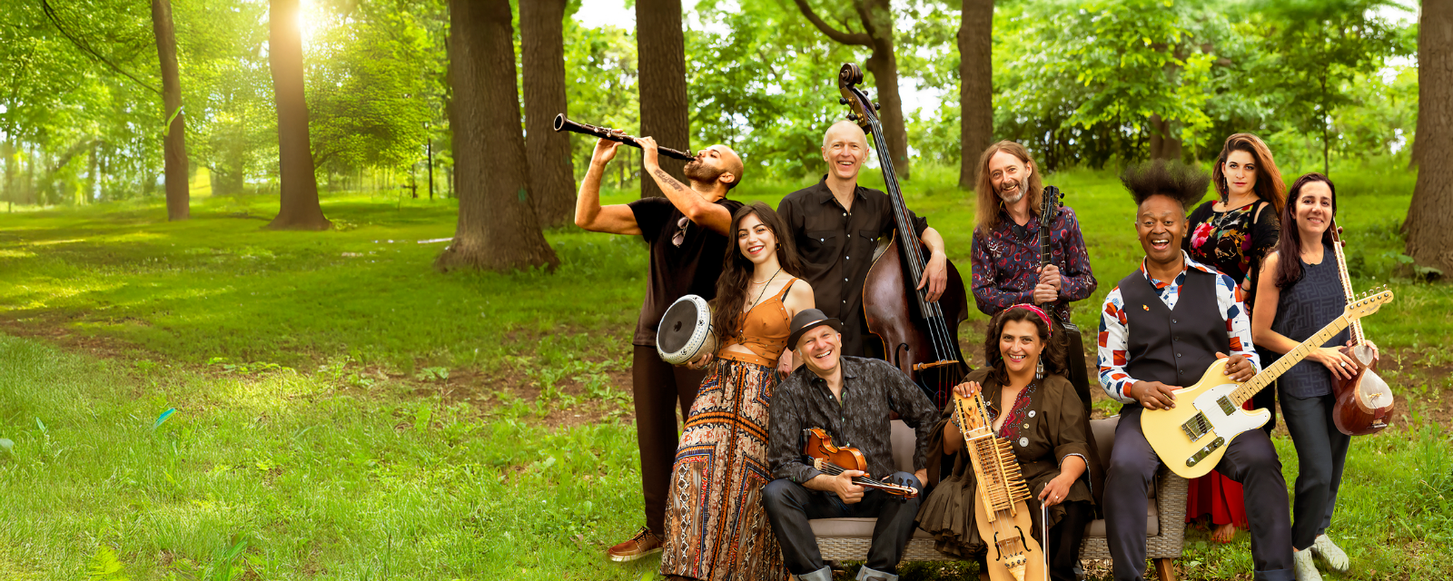 Group of musicians sits outside in green space on a bright sunny day