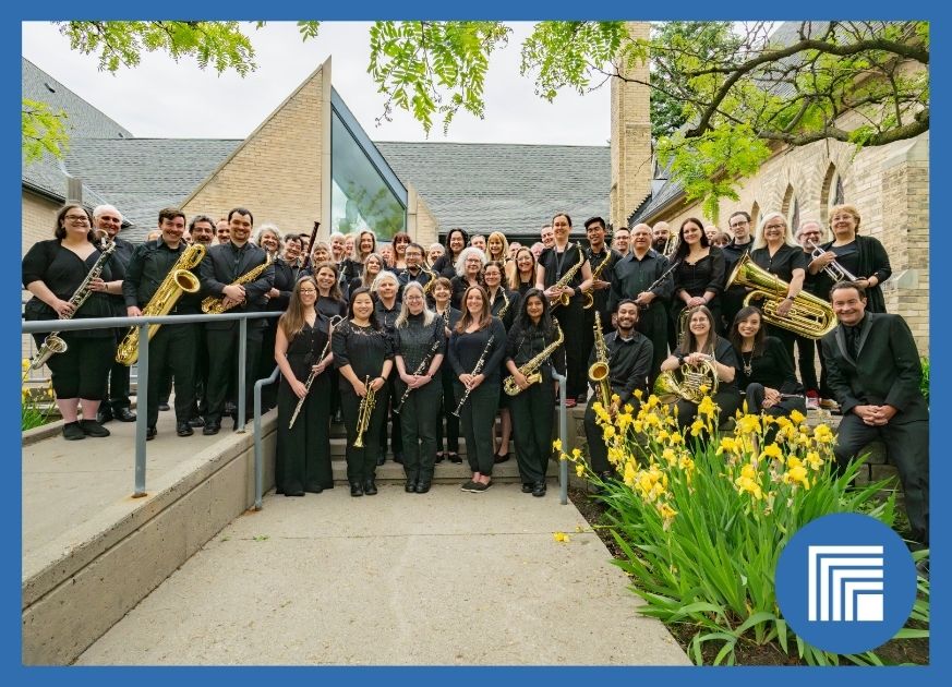 A large instrumental band holding their instruments in front of a building during the summer months